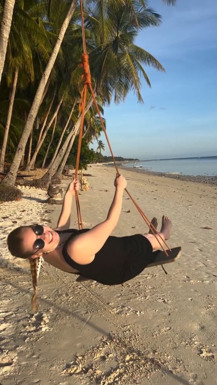 Frau mit Sonnenbrille sitzt lächelnd auf einer Schaukel am Strand